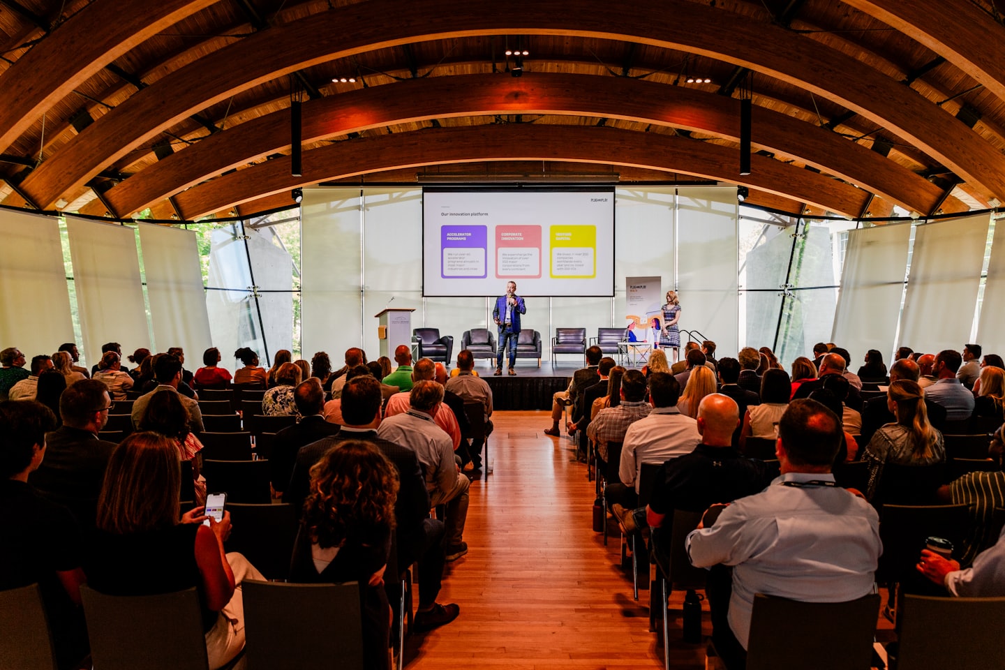 Speakers present to a large audience inside a wooden auditorium.